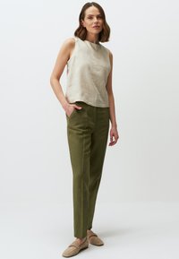 Beige Leinen-Hemdtopp ohne Ärmel mit rundem Ausschnitt, kombiniert mit grünen, geraden Hosen. Das Model trägt hellbeige Slipper.