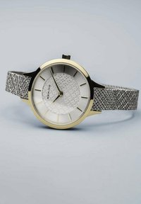 Round silver watch with a mesh band, gold accents, textured white dial, and minimalist hour markers. The case has a sleek black finish.