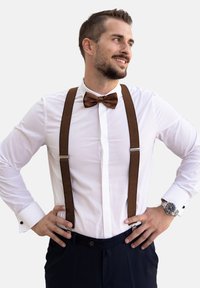 Una pajarita marrón y tirantes acentúan una camisa blanca. El hombre lleva pantalones azul marino y un reloj, mostrando un aspecto formal pulido.
