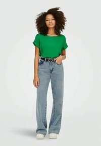 Donna con capelli ricci che indossa una maglietta verde a maniche corte, jeans a vita alta con gamba larga, cintura nera e sneakers bianche, in piedi con una mano in tasca.