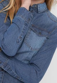 Person som bär en knappad långärmad denimskjorta med en bröstficka, vilar handen nära halsen, beskuren från hakan och neråt.