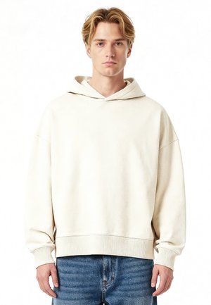 Jeune homme aux cheveux blonds portant un sweat à capuche beige clair surdimensionné et un jean bleu, debout devant un fond blanc.