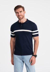 Mörkblå kortärmad polo skjorta med vita horisontella randar på ärmarna och bröstet, tillverkad av ett stickat tyg, i kombination med ljusblå jeans.