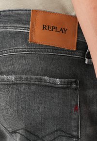 Grey denim jeans featuring a leather label with "REPLAY" embossed. Notable stitching details and a small red "R" on the back pocket.