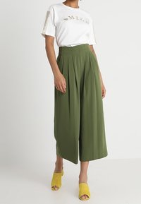 Nainen, joka pukeutuu valkoiseen "SMILE"-t-paitaan, korkeavyötäröisiin oliivinvihreisiin leveälahkeisiin housuihin ja keltaisiin avonaisiin korkokenkiin, seisoo tylsällä taustalla.