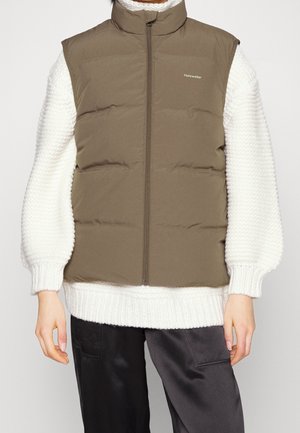 Persona que lleva un chaleco acolchado marrón sobre un suéter blanco de punto con pantalones negros, de pie frente a un fondo claro y liso.