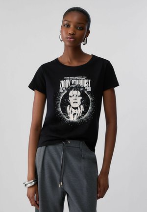 Donna con t-shirt nera con grafica Ziggy Stardust, pantaloni grigi con coulisse e orecchini e braccialetti a cerchio in argento, in piedi davanti a uno sfondo semplice.