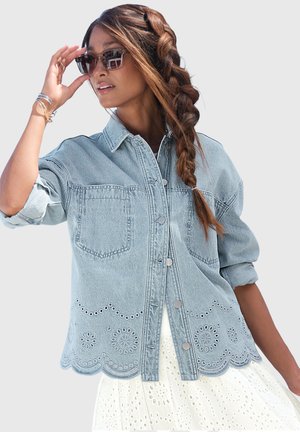 Woman wearing light blue denim jacket with eyelet embroidery, sunglasses, and white eyelet dress, holding sunglasses with braided hair over shoulder.