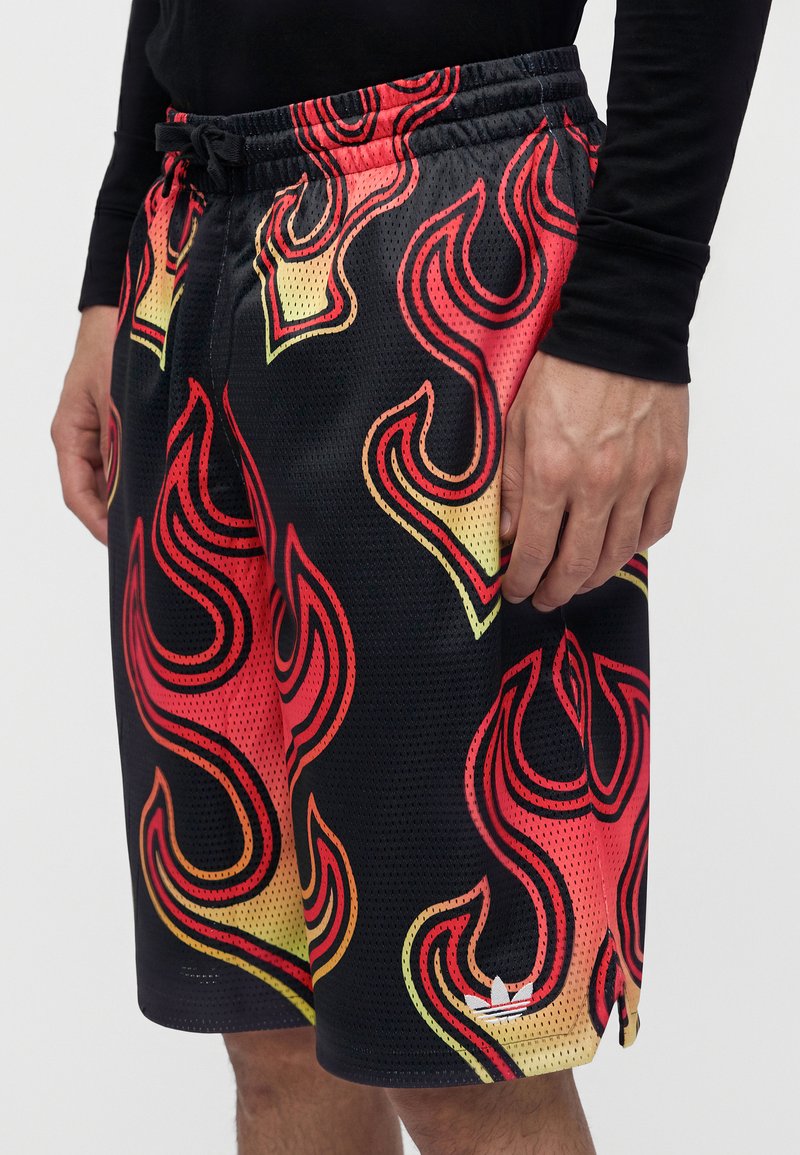 Uomo che indossa pantaloncini in rete neri con motivi di fiamme rosse, arancioni e gialle, e una maglia nera a maniche lunghe, in piedi con una mano visibile.