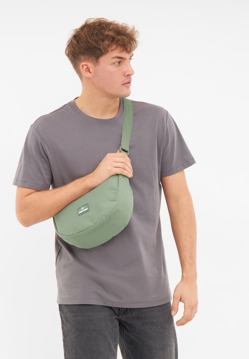 Borsa a spalla di tessuto verde con forma arrotondata, tracolla regolabile e un piccolo logo sul fronte, tenuta da una persona che indossa una maglietta grigia.