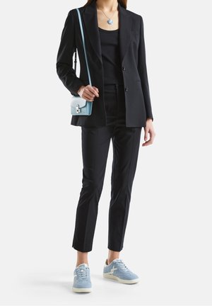 Frau trägt einen schwarzen Blazer und eine schwarze Hose, ein schwarzes Oberteil, blaue Sneakers, eine blaue Umhängetasche und eine runde blaue Anhängerkette.