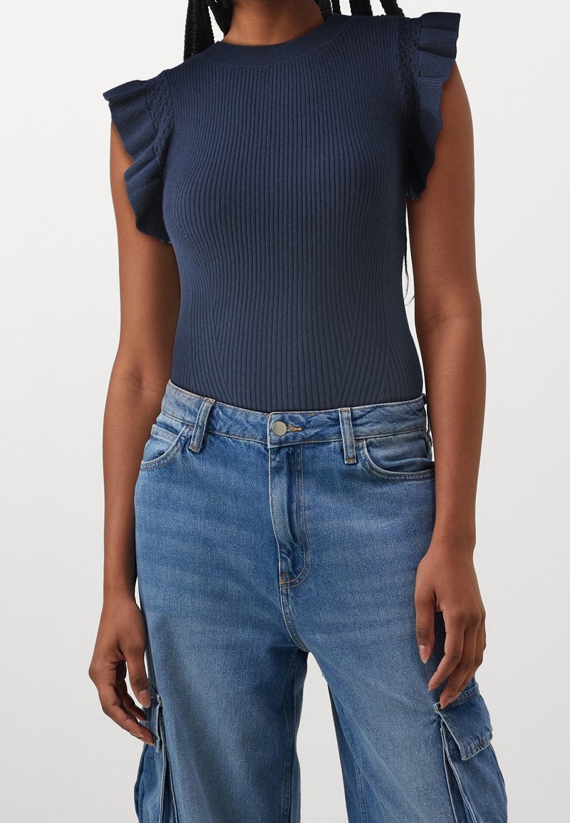 Femme portant un top sans manches côtelé bleu marine avec des épaules volantées et un jean cargo taille haute bleu clair.