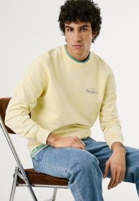 Gele sweatshirt met geribbelde manchetten, met een subtiele logodetail. Gelaagd over een blauw-groene shirt, gecombineerd met lichte spijkerbroek.