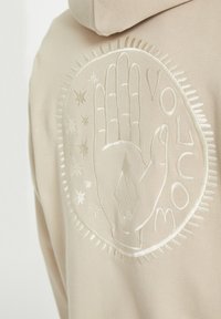 Sweat à capuche beige avec un motif en relief représentant une main ouverte, des étoiles et le mot « VOLCOM » disposés en cercle dans le dos.