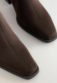 Brown suede ankle boots with a pointed toe design and a flat black sole. Features stitched detailing along the sides.
