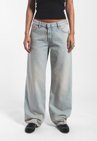 Hellblaue, weit geschnittene Jeans aus Denim, mit hoher Taille, Knopfverschluss und Spitzenstickerei. Getragen mit einem figurbetonten schwarzen Crop-Top.