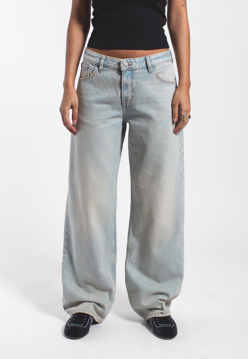 Hellblaue, weit geschnittene Jeans aus Denim, mit hoher Taille, Knopfverschluss und Spitzenstickerei. Getragen mit einem figurbetonten schwarzen Crop-Top.