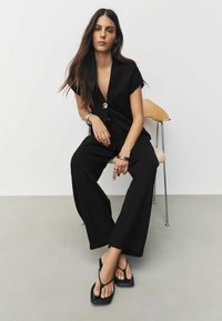 Schwarzes Outfit mit tiefem V-Ausschnitt, kurzen Ärmeln und weitem Hosenbein. Accessoires sind silberne Armbänder und schwarze Flip-Flops.