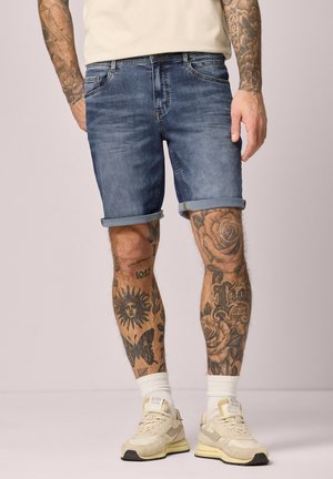 Homme portant un short en jean retroussé, des chaussettes blanches, des baskets beiges, et exhibant des tatouages de soleil, papillon, roses et texte sur les jambes et les bras.