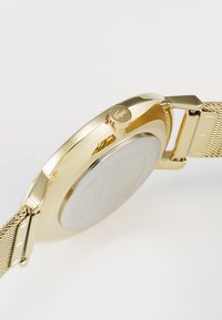 Montre dorée avec un bracelet en maille, boîtier circulaire et un fond rond en argent. Présente une couronne latérale saillante avec un accent de logo.