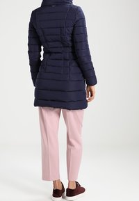 Námornická péřová bunda s horizontálním prošíváním, vysokým límcem a přizpůsobeným střihem, kombinována s světle růžovými kalhotami a burgundskými botami.