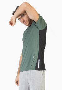 Camiseta deportiva verde con panel lateral negro, que presenta un logo y un bolsillo con cremallera. Textura suave, mangas cortas y un corte relajado.