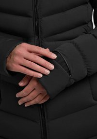 Giacca a scomparsa nera con design trapuntato, apertura con zip e polsini elasticizzati. Il materiale ha una texture liscia con una finitura lucida.