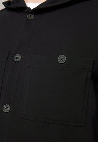 Giacca nera a collo alto con cappuccio, design strutturato, tessuto testurizzato e tasca sul petto con bottoni coordinati.