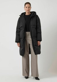 Giacca a piumino nera con cappuccio, dotata di chiusura con zip, due tasche frontali e tessuto trapuntato. Indossata sopra una maglia nera e pantaloni beige.