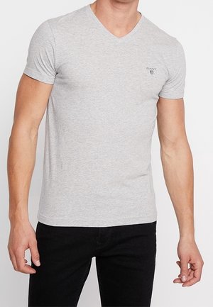 Mężczyzna noszący dopasowany jasnoszary t-shirt z dekoltem w serek i małym logo na piersi, w połączeniu z czarnymi spodniami, stoi na jednolitym tle.