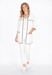 Cardigan alb cu margini și buzunare negre, mâneci trei sferturi, asociat cu pantaloni albi și teniși. Textură netedă, croială lejeră.