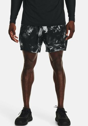 Mand iført sorte og grå mønstrede sportsshorts, sort langærmet trøje og sorte løbesko, stående med fødderne lidt fra hinanden.