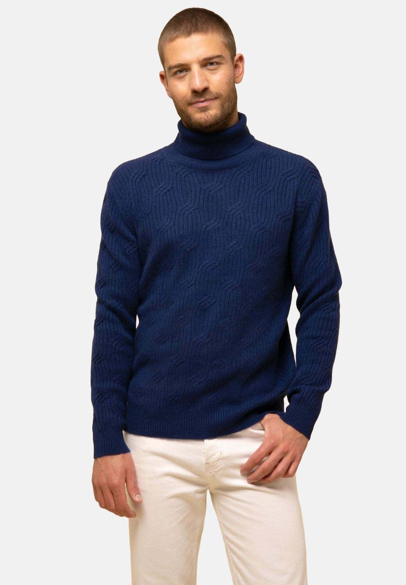 Jersey de cuello alto azul marino con un patrón de punto texturizado en forma de ola. Puños y dobladillo acanalados, material suave, combinado con pantalones de color claro.