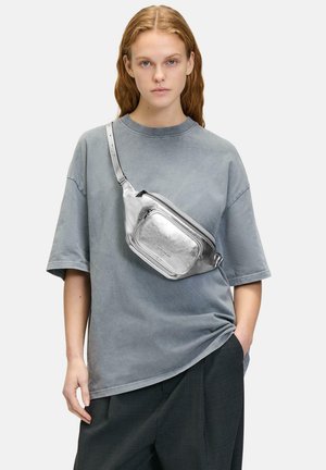 Silberfarbige metallische Crossbody-Tasche mit Reißverschluss, rechteckiger Form, verstellbarem Tragegurt und glatter Textur, getragen zu einem übergroßen grauen T-Shirt.