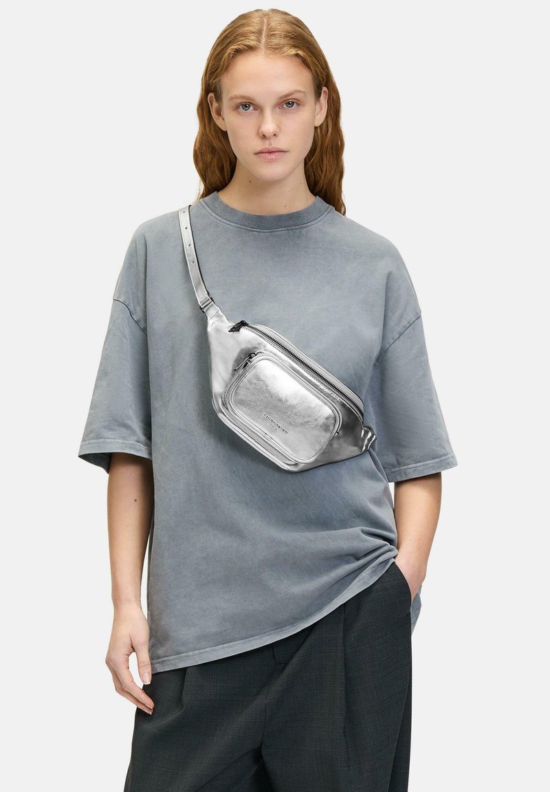 Srebrna metaliczna torebka na ramię z zamkiem błyskawicznym, prostokątny kształt, regulowany pasek i gładka faktura, noszona z oversize'owym szarym T-shirtem.