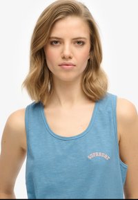 Niebieski top na ramiączkach wykonany z lekkiego materiału, z okrągłym dekoltem i wyhaftowanym logo "Superdry" w różowym kolorze nad klatką piersiową.