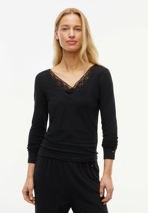 Femme blonde portant un haut noir à manches longues avec un détail en dentelle au col en V, associé à un pantalon noir sur fond blanc.