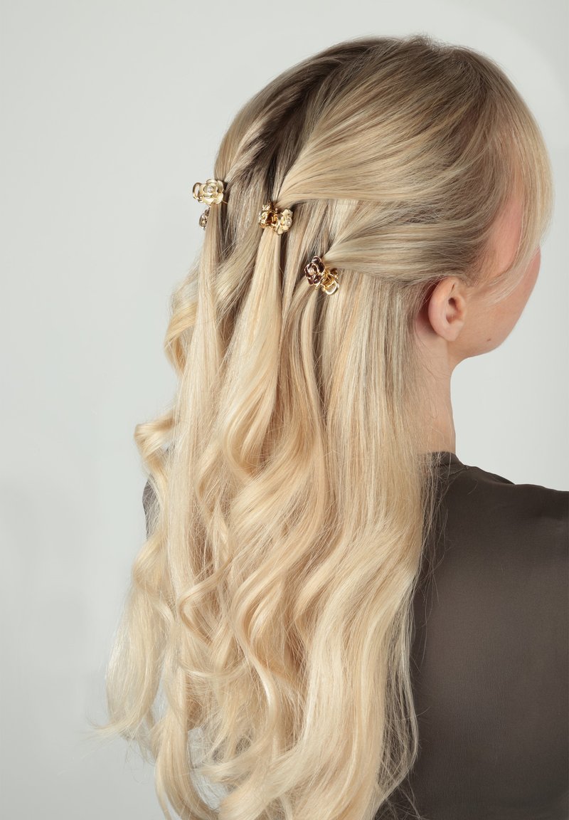 Cheveux blonds coiffés en boucles lâches, partiellement maintenus en arrière avec trois barrettes florales dorées, portant un haut sombre, légèrement tourné vers la droite.