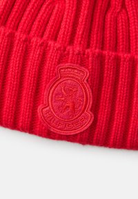 Czerwona dzianinowa czapka beanie o ryflowanej strukturze. Posiada wypukłą, okrągłą łatę z logo przedstawiającym lwa oraz napisem "HILFIGER" z przodu.