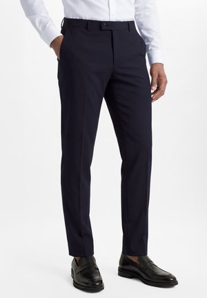 Man draagt op maat gemaakte marineblauwe pantalon, wit overhemd, zwarte leren loafers, met handen in de zakken tegen een effen achtergrond.