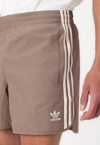 Rudos lengvi šortai su trimis baltomis juostelėmis šoninėse dalyse ir išsiuvinėtu Adidas logotipu apatinėje dalyje. Elastingas juosmens juostos detalė.