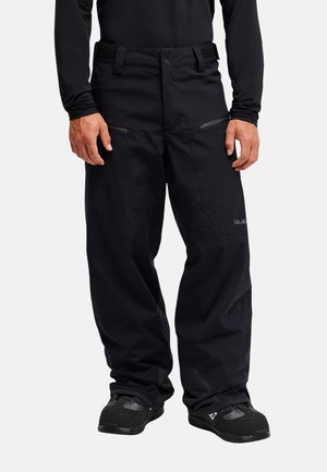 Mann trägt schwarze Outdoor-Hose mit Reißverschlusstaschen und schwarzen Schuhen, steht vor weißem Hintergrund.