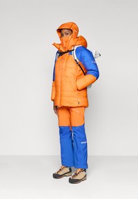 Jachetă izolată cu blocuri de culoare portocalie și albastră, dotată cu glugă și închidere cu fermoar. Asortată cu pantaloni albaștri și portocalii și bocanci de drumeție bej.