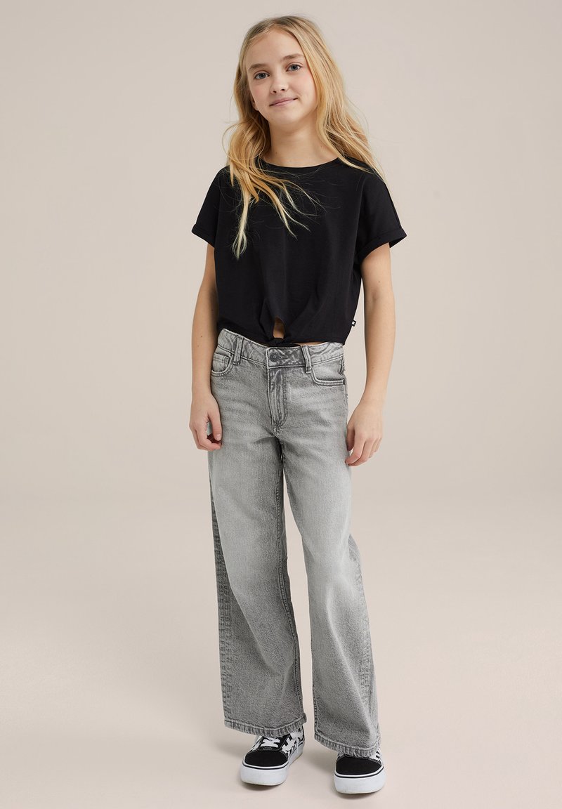 Sort T-shirt med korte ærmer og knude detalje foran, parret med lysegrå brede jeans og sorte sneakers med hvide accenter.