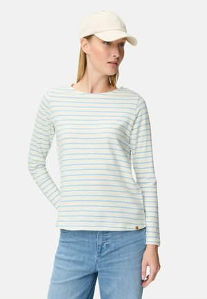 Vrouw met een wit shirt met lange mouwen en lichtblauwe horizontale strepen, lichtblauwe spijkerbroek en een crèmekleurige baseballpet.