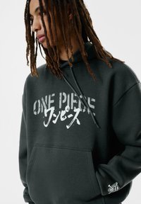 Sötétzöld kapucnis pulóver, puha anyagból, fehér színű 'ONE PIECE' felirattal és egy kis logóval az ujjakon. Elöl zseb található.