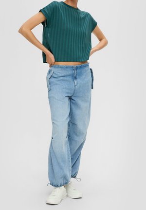 Personne portant un jean bleu clair ample avec des poignets élastiques, des baskets blanches et un crop top vert rayé à manches courtes sur un fond uni.