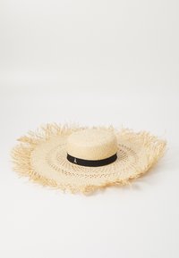 Laaja lieriöinen olkihattu, jossa on fringed-reuna, valkoisella kiinteällä nauhalla ja kultaisella emblemilla. Luonnollinen väri ja kudottu rakenne.