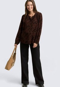 Blouse à imprimé léopard marron et noir avec manches longues, détail à boutons au niveau de l'encolure, associée à un pantalon large noir et un sac à main beige.
