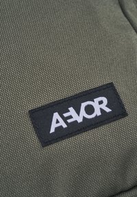 Green textured fabric with a black rectangular patch displaying the white label "A-E-VOR." Close-up view highlights material details.